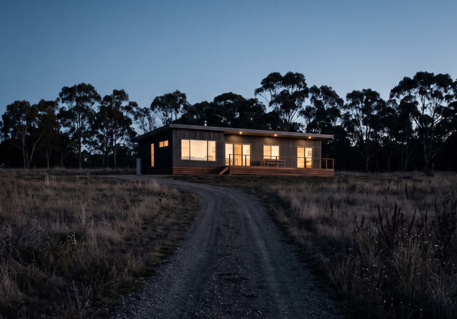 A single-storey modular home with lit windows at dusk, at the end of a gravel driveway on a rural block